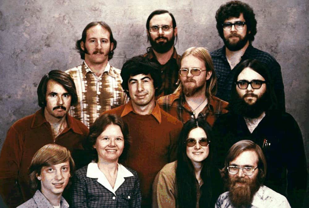 Foto antiga em tons sépia de um grupo de doze pessoas posando juntas em três fileiras, com cabelos longos, bigodes e óculos, em estilo dos anos 1970