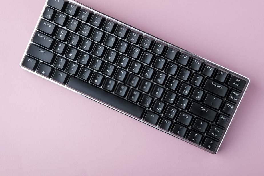 Teclado mecânico preto compacto com teclas em caracteres cirílicos e latinos sobre fundo rosa claro, visto de cima