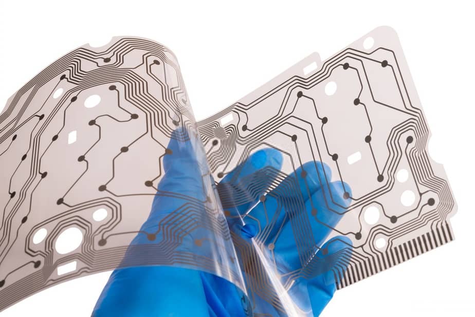 Mão com luva azul segurando uma membrana transparente de circuito impresso com trilhas prateadas e contatos, sobre fundo branco