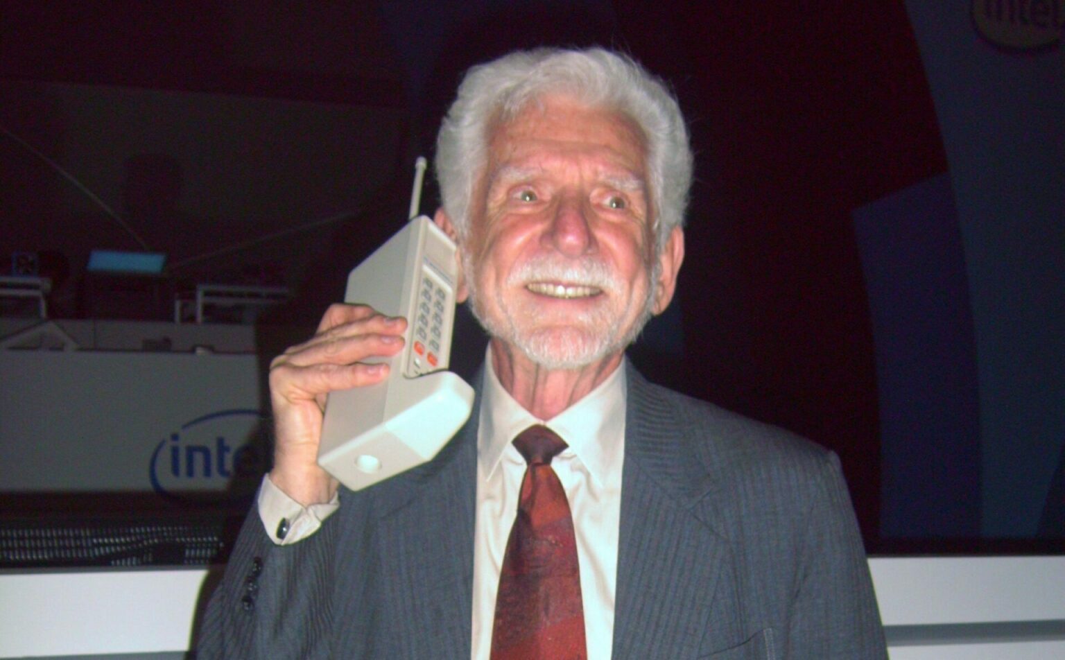 Homem de cabelos brancos e terno cinza segura um celular antigo, grande e de teclado, encostado na orelha, com logo da Intel ao fundo