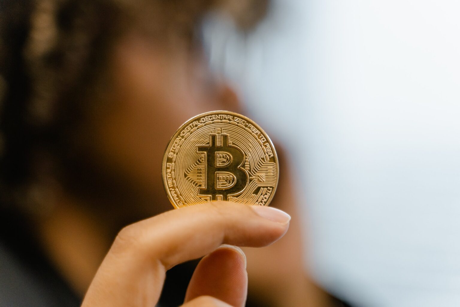 Moeda dourada com o símbolo do Bitcoin segurada entre os dedos de uma pessoa, com fundo desfocado