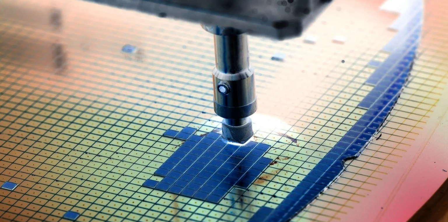 Wafer de silício com grade de chips em processo de fabricação, com sonda metálica de teste tocando um dos dies, em ambiente de fábrica de semicondutores