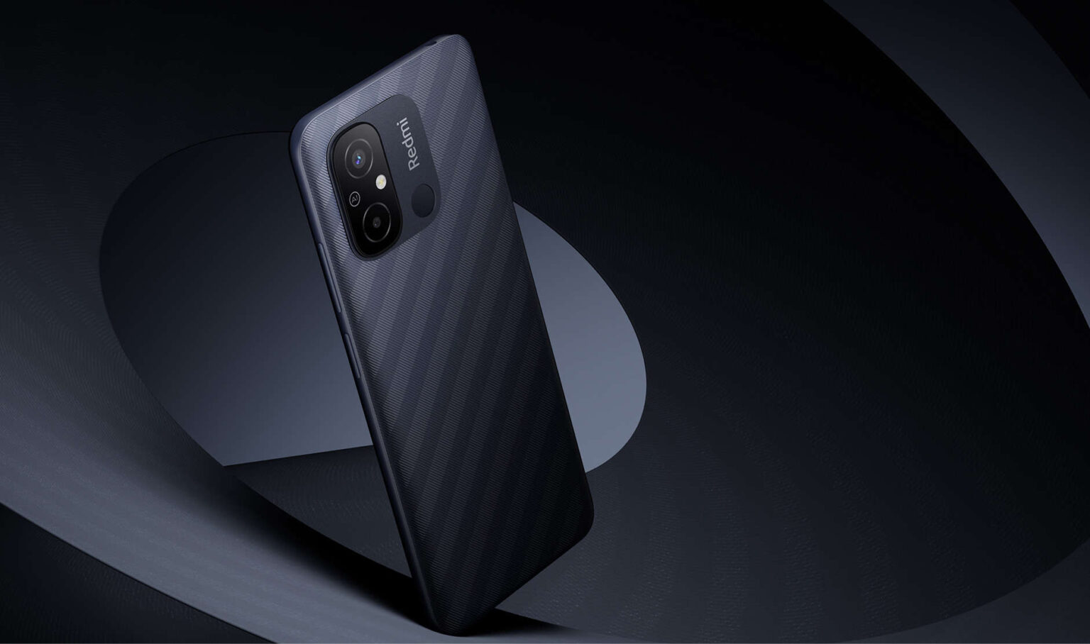 Smartphone Redmi 12C de cor preta com textura diagonal na traseira, câmeras e sensor de digital, apoiado em fundo escuro com formas geométricas