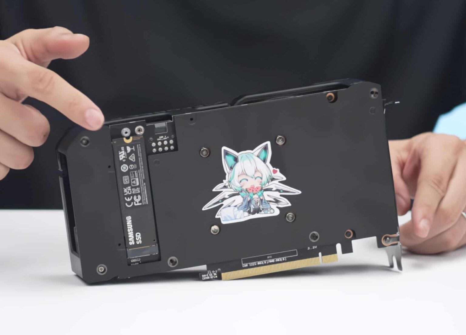 Verso de placa de vídeo preta com um SSD Samsung M.2 instalado sobre o PCB e um adesivo de personagem de anime, com mãos apontando para o conjunto