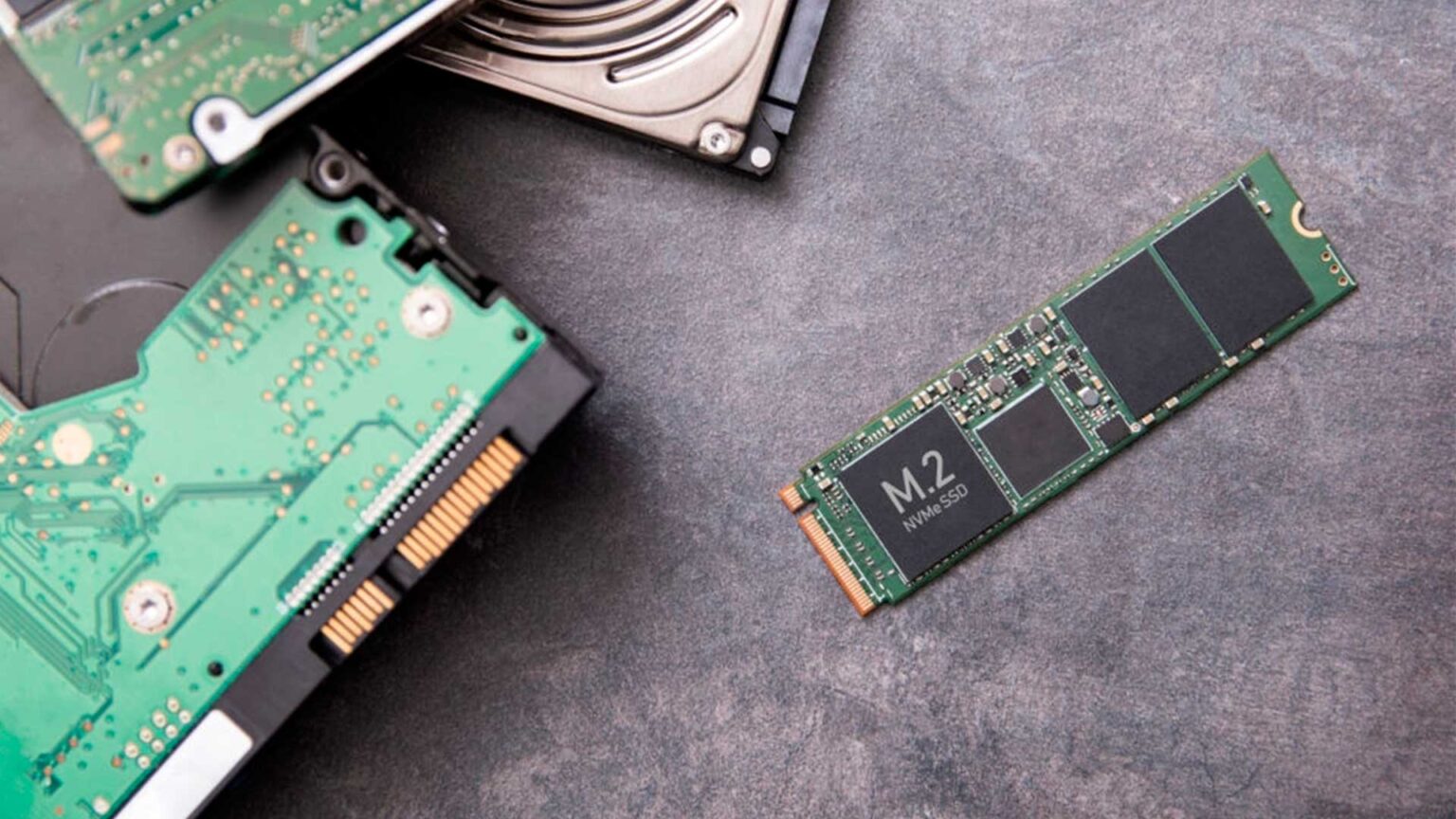 SSD M.2 NVMe ao lado de um HD mecânico de 3,5 polegadas com placa de circuito verde, sobre uma superfície cinza