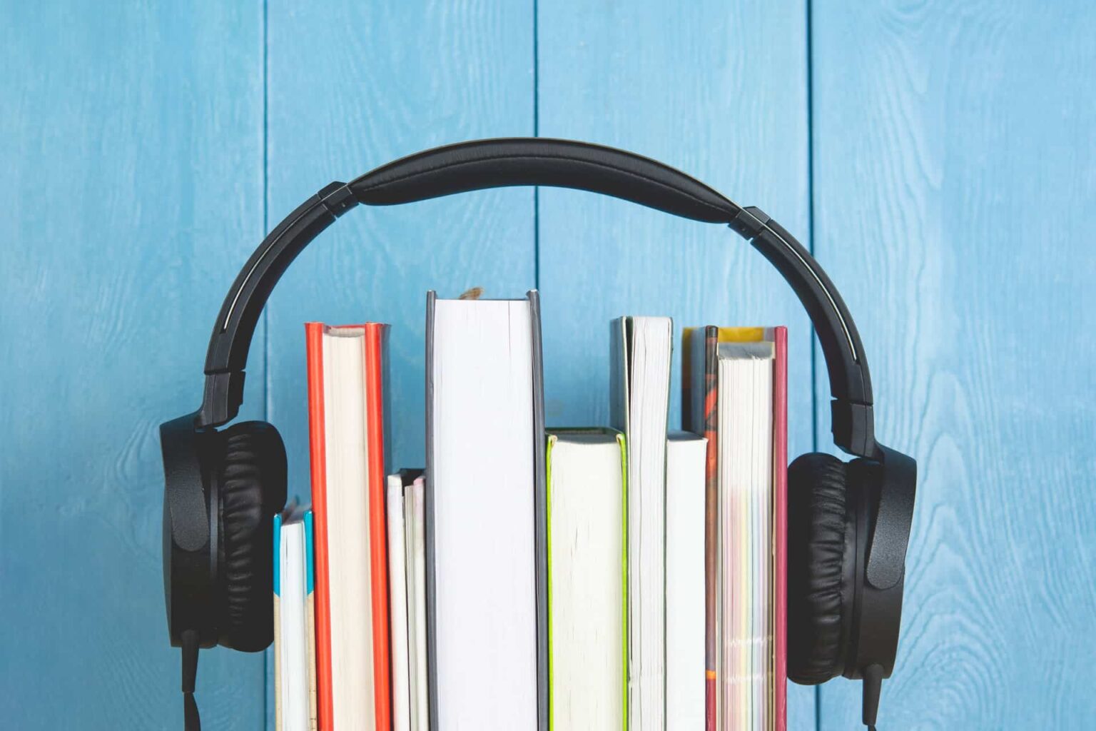 Headphones pretos apoiados sobre uma fileira de livros de capa dura, em frente a um fundo azul de madeira
