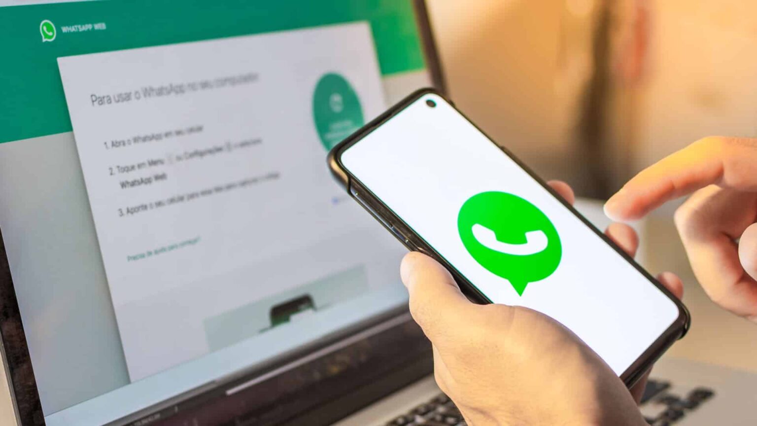 Mão segurando smartphone com logo do WhatsApp na tela, ao lado de notebook exibindo a página do WhatsApp Web com instruções de uso