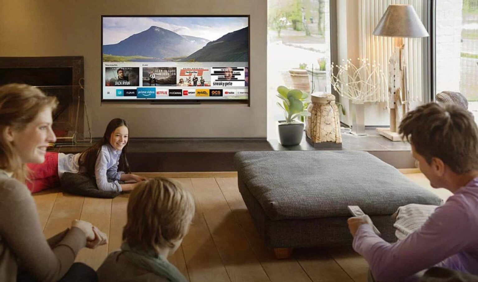 Família sentada no chão de uma sala assistindo a uma smart TV que exibe menu de streaming com paisagem de montanhas ao fundo