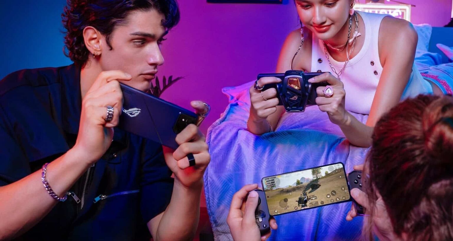 Três pessoas jogando em smartphones com controles periféricos encaixados, sob luz roxa e azul, em um celular ASUS ROG com jogo de tiro em tela