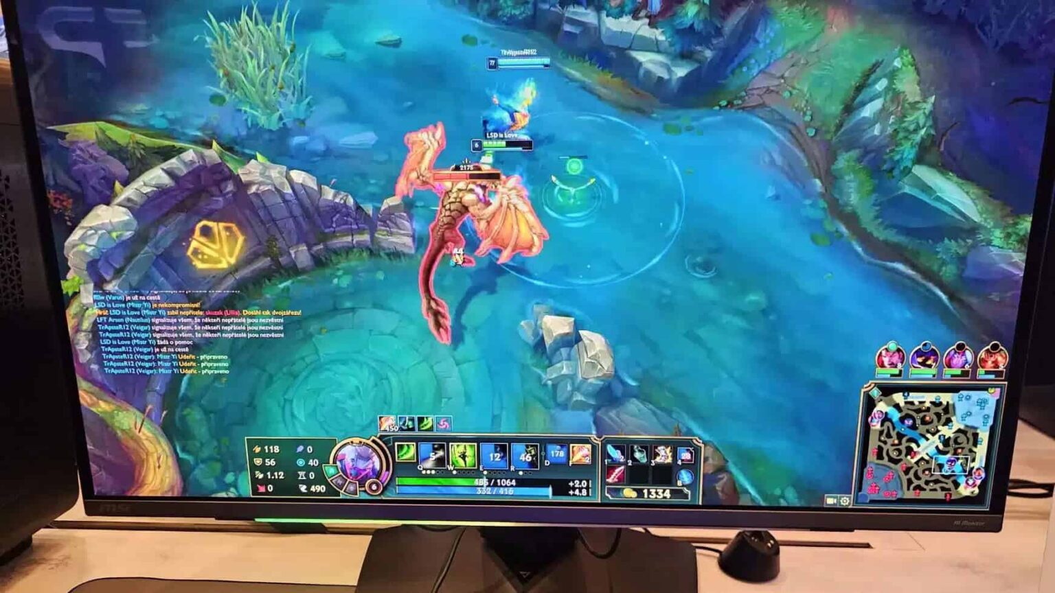 Monitor exibindo partida de League of Legends com personagem serpentino em rio azul, chat de jogo e HUD com vida, mana e mapa, sobre mesa com mouse