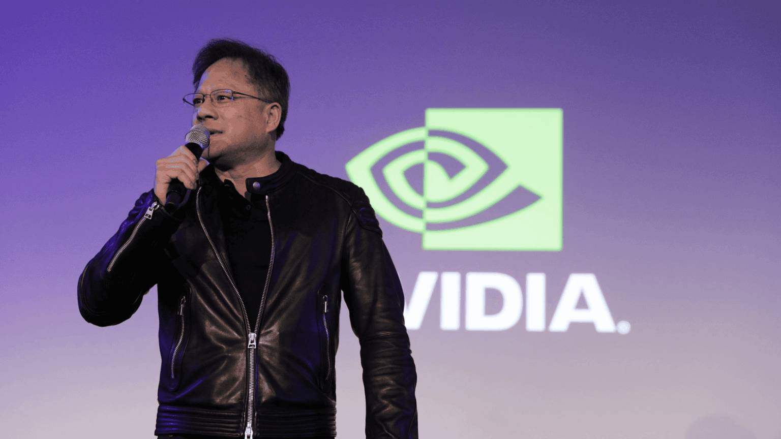 Homem de jaqueta de couro preto falando ao microfone em palco, com o logotipo da NVIDIA ao fundo em tons roxos