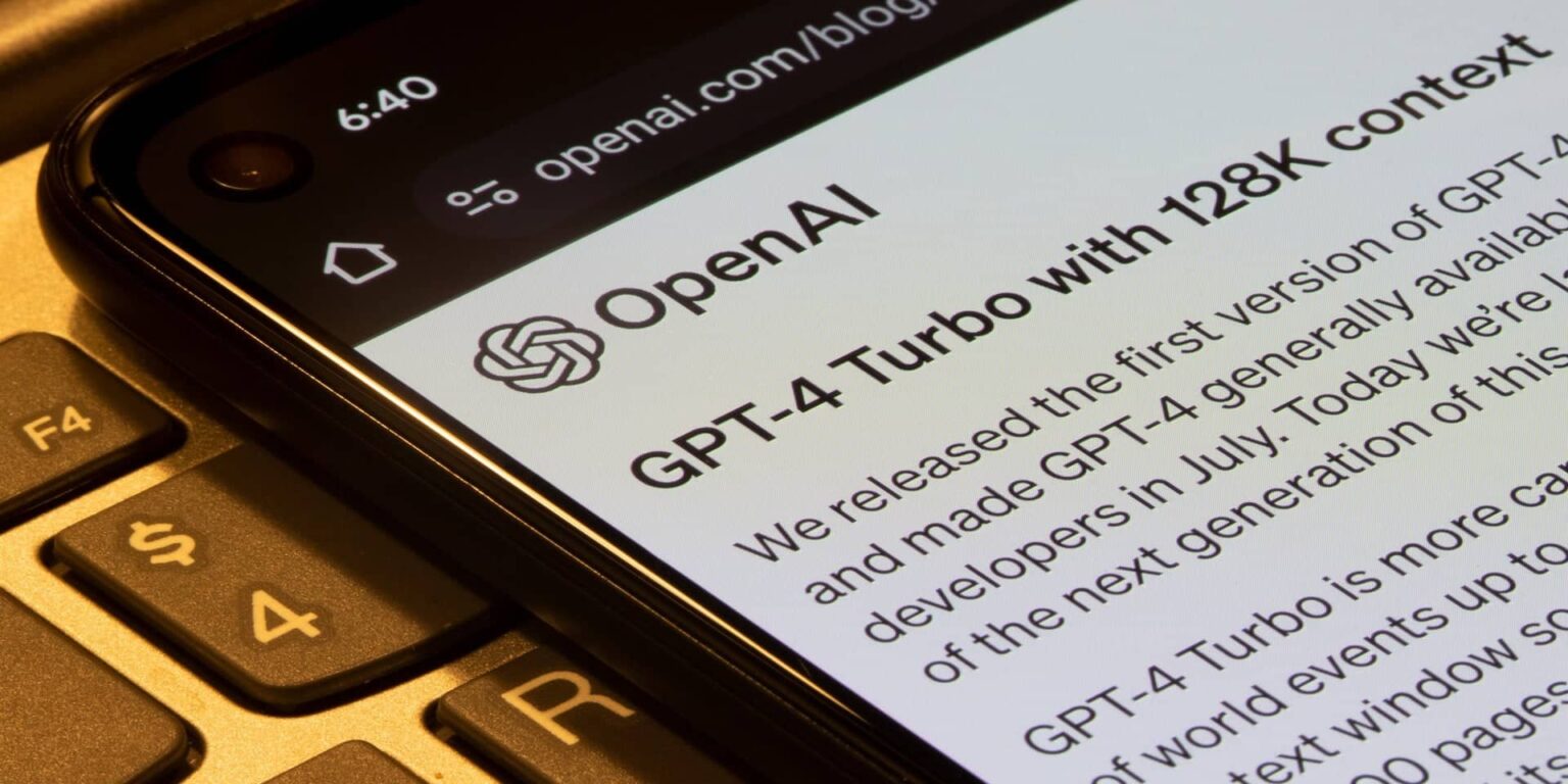 Smartphone exibindo o blog da OpenAI com o anúncio do GPT-4 Turbo com contexto de 128K, sobre um teclado de notebook