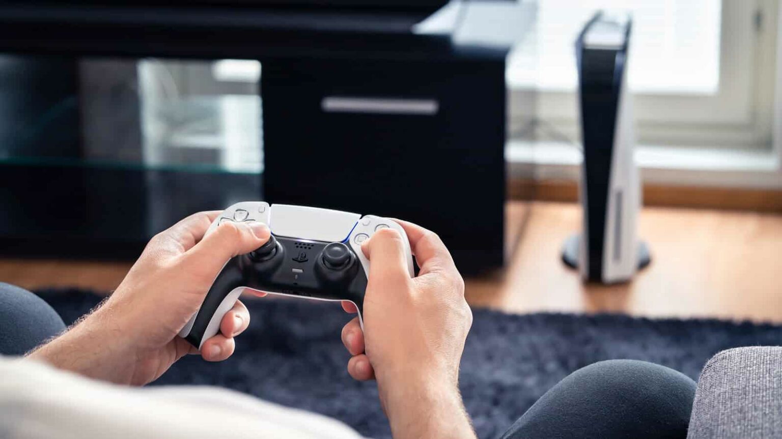 Pessoa segura controle DualSense branco do PlayStation 5 em sala, com console PS5 ao fundo sobre a estante