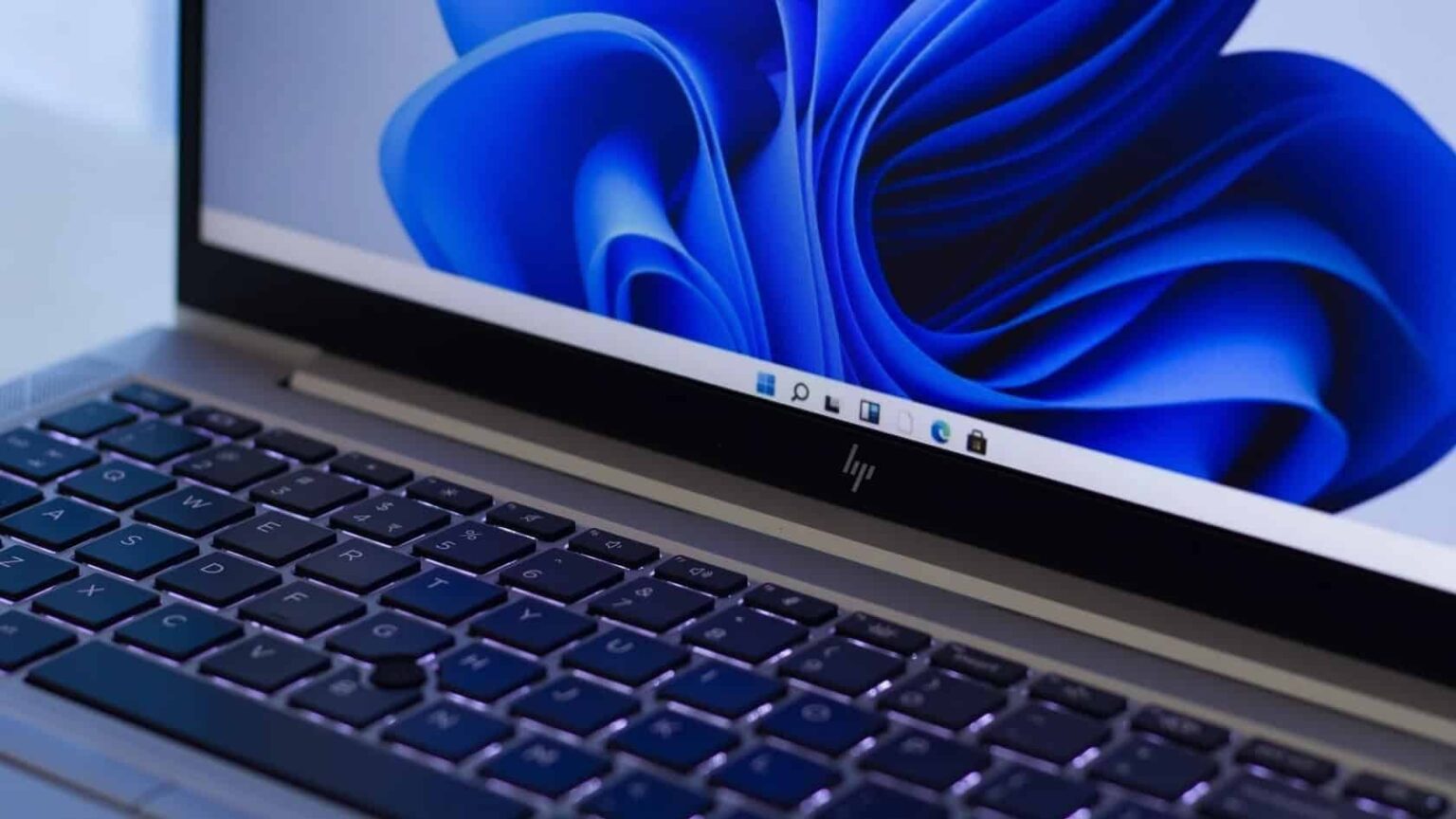 Notebook HP com papel de parede azul em tela do Windows 11 e teclado iluminado, com a barra de tarefas visível na parte inferior da tela