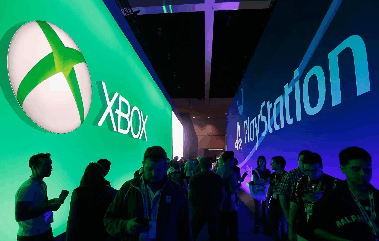 Pessoas circulam em evento de games entre painéis iluminados com as marcas Xbox, à esquerda em verde, e PlayStation, à direita em azul
