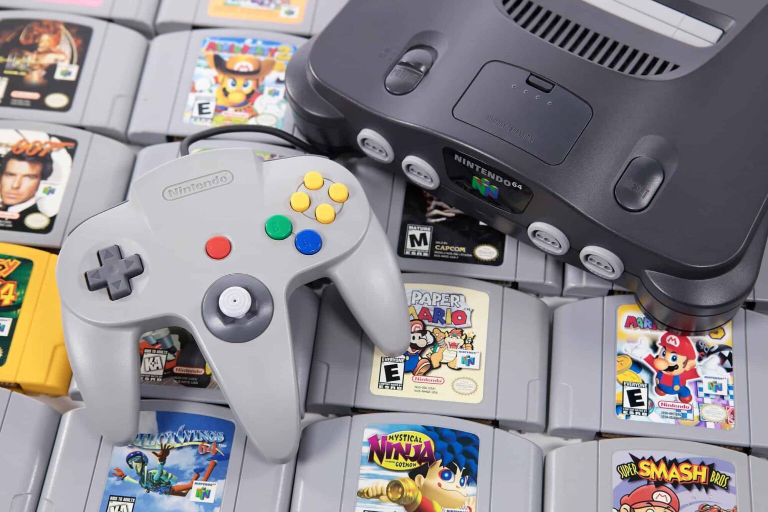 Console Nintendo 64 cinza com controle cinza sobre uma pilha de cartuchos de jogos como Paper Mario, Super Smash Bros. e Mystical Ninja