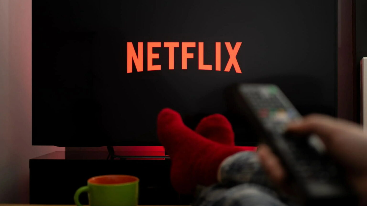 Televisão com o logo da Netflix na tela, pés de meia vermelha apoiados em uma mesa, caneca verde e controle remoto em primeiro plano