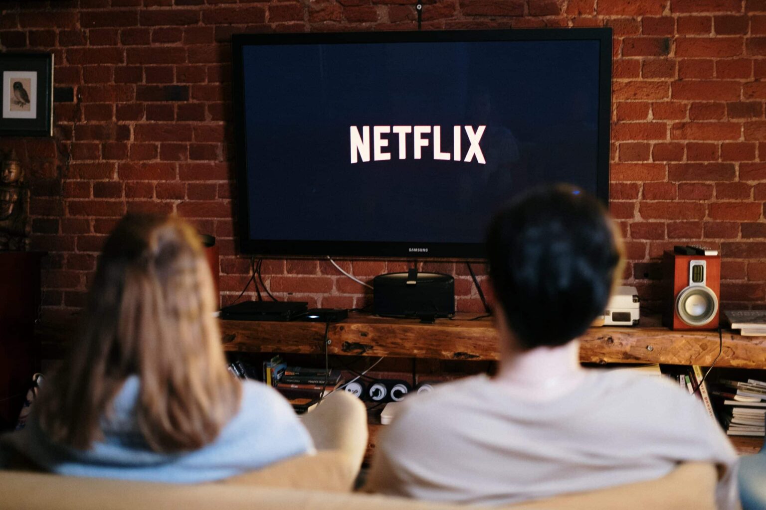 Duas pessoas de costas assistem à logo da Netflix em uma TV grande sobre um móvel de madeira, em uma sala com parede de tijolos