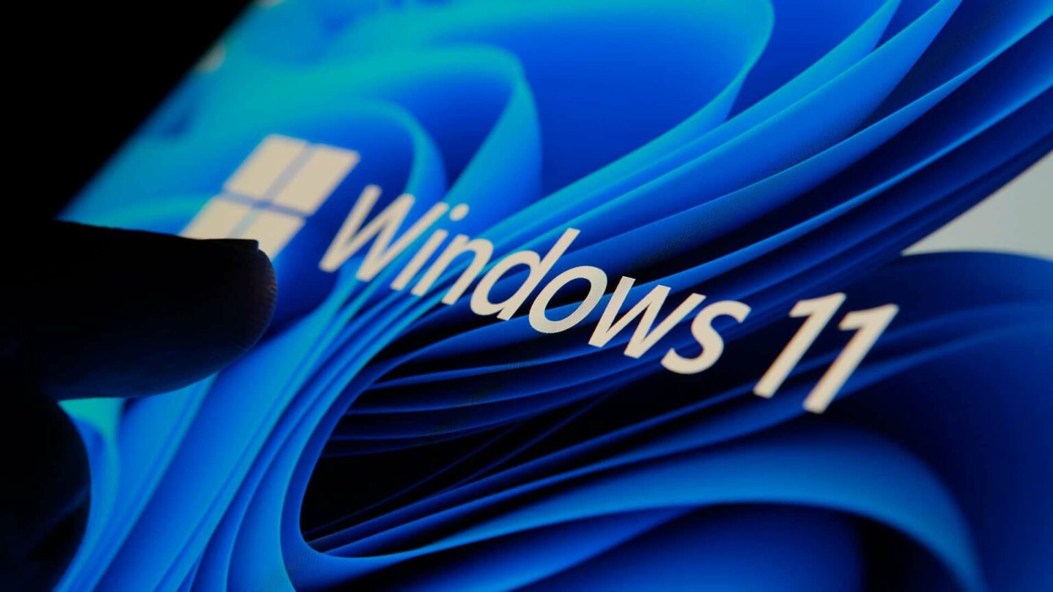 Logo do Windows 11 em branco sobre fundo azul com curvas, com a sombra de um dedo tocando a tela no canto esquerdo