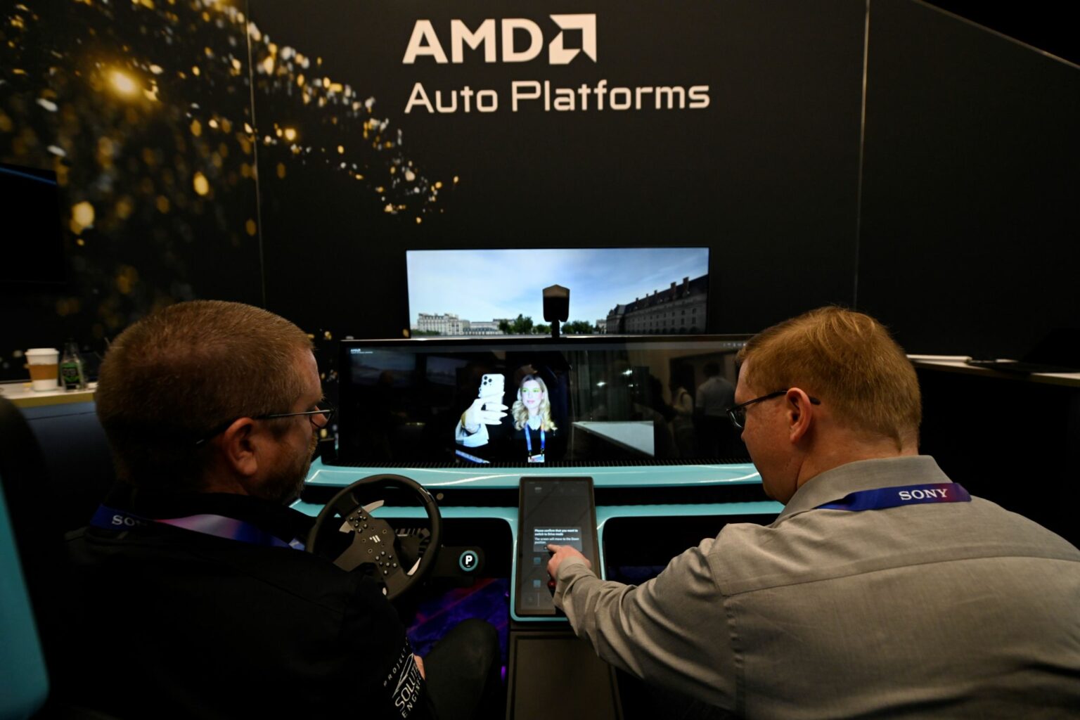 Dois homens em simulador de carro com volante, diante de painel com tela, exibindo a plataforma AMD Auto Platforms em evento