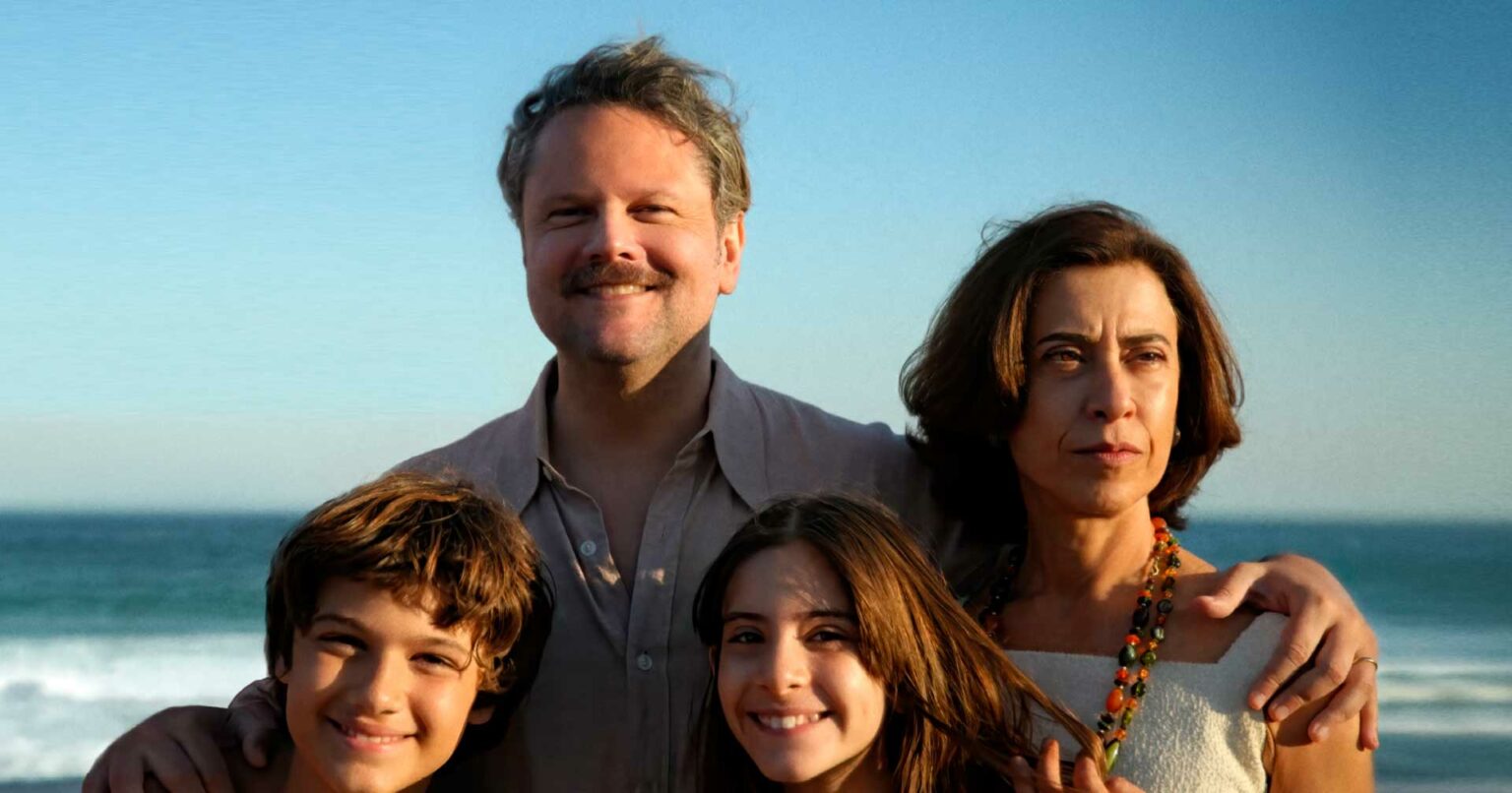 Família reunida na praia com um homem de camisa bege atrás, uma mulher de colar colorido e duas crianças sorrindo em primeiro plano, com o mar ao fundo