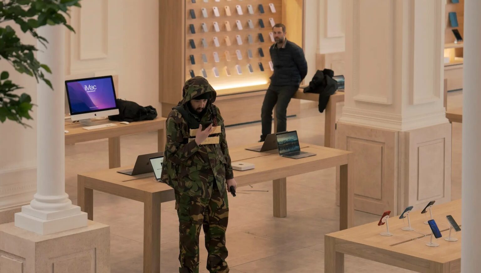 Loja com iMac e MacBooks expostos; homem de camuflagem com capuz caminha entre as mesas, enquanto outro homem observa ao fundo