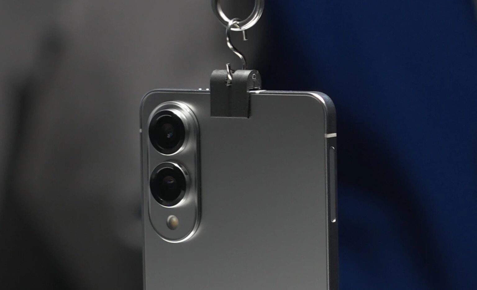 Traseira de smartphone prata fino com dois sensores de câmera e flash, com um gancho metálico preso à borda superior
