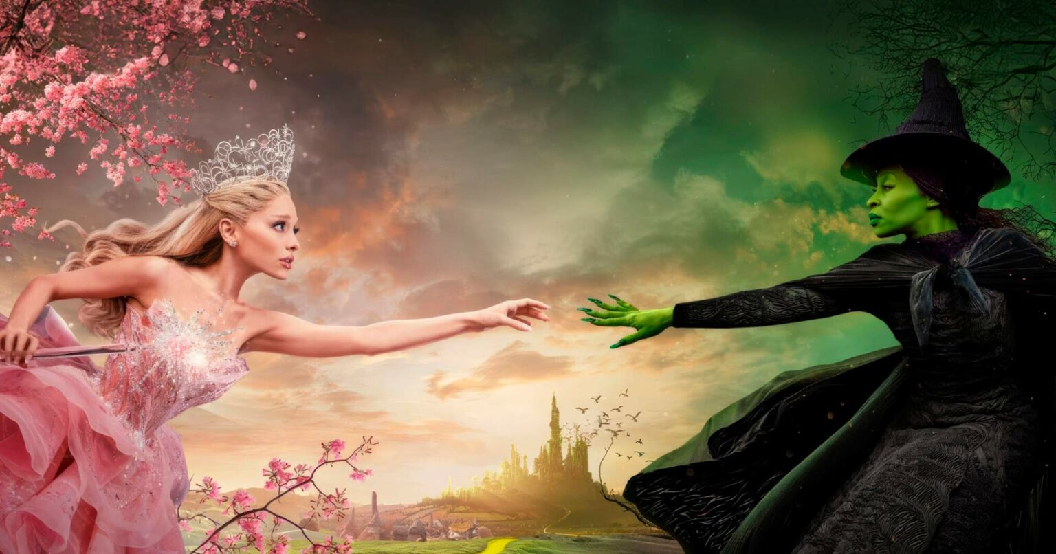 Pôster promocional do filme Wicked com a Glinda de vestido rosa e coroa, estendendo a varinha, e a Bruxa Má do Oeste de chapéu preto e pele verde, com a cidade…