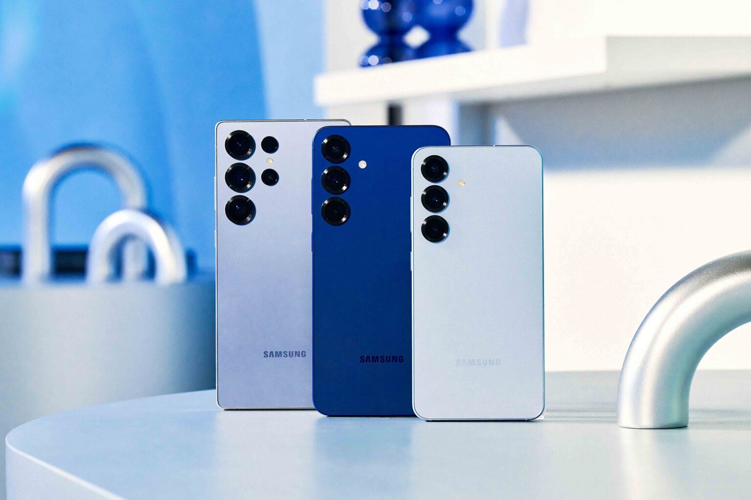 Três smartphones Samsung Galaxy S lado a lado, de cores cinza, azul e branca, vistos pela traseira sobre uma mesa clara