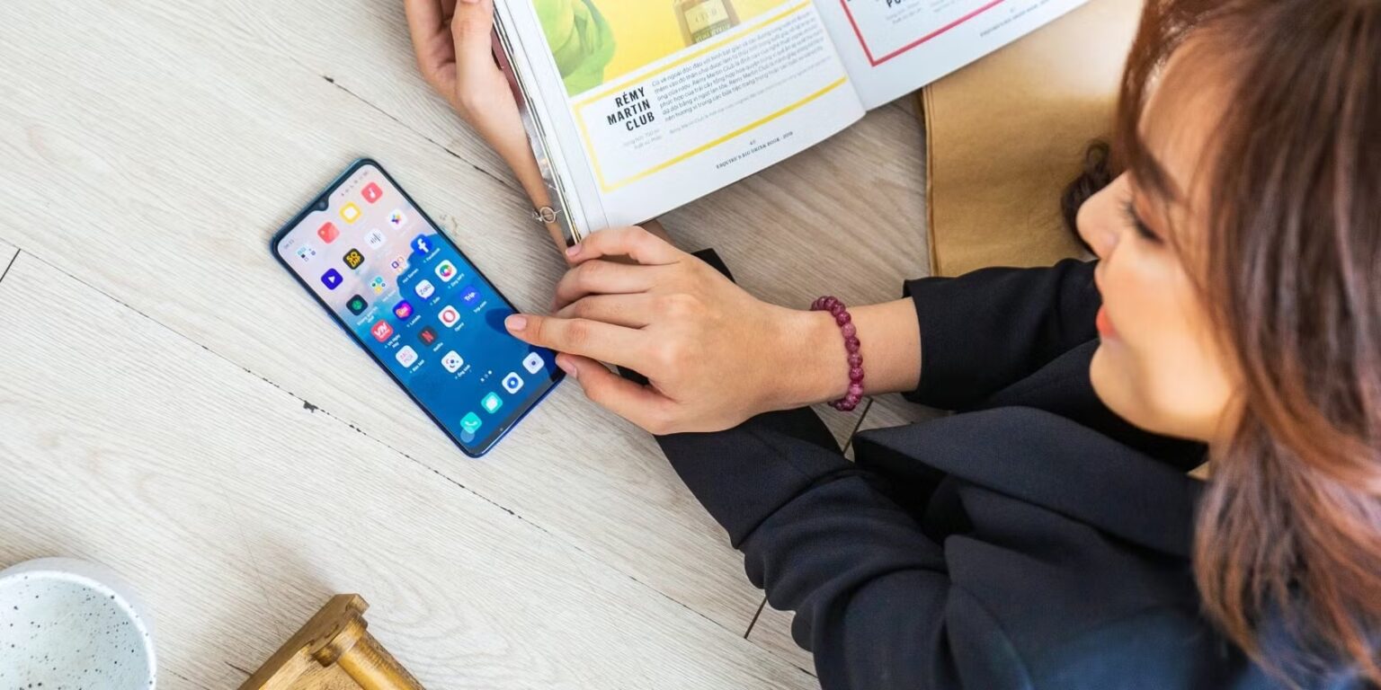 Mulher de blusa escura lê revista enquanto toca na tela de um smartphone Android com ícones de aplicativos sobre uma mesa de madeira clara