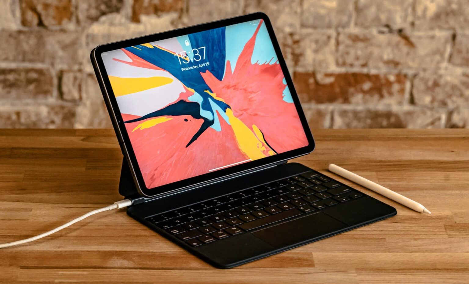 iPad Pro com teclado Magic Keyboard e Apple Pencil sobre mesa de madeira, exibindo papel de parede colorido