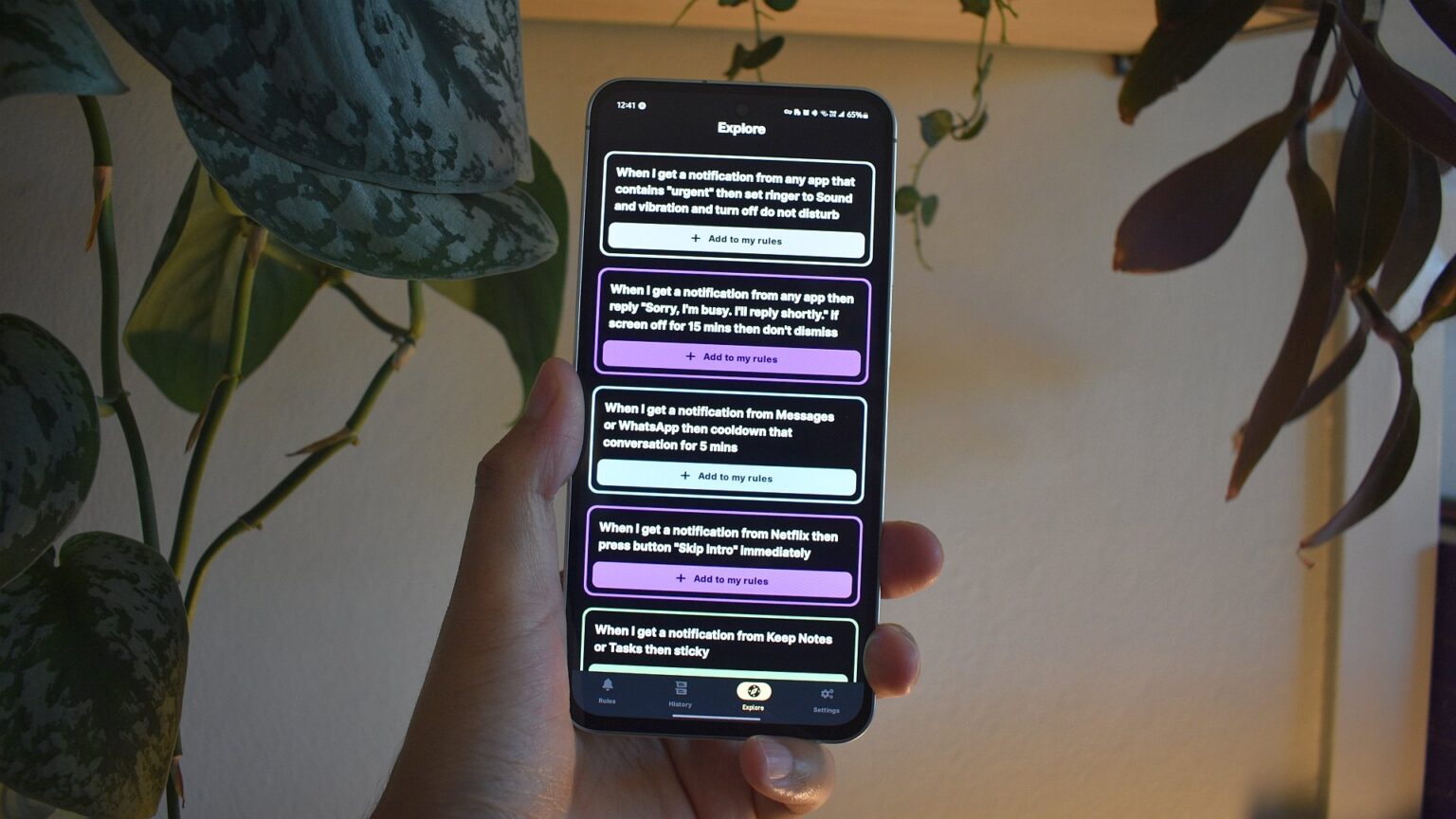 Mão segurando smartphone exibindo a tela Explore de um app de automação, com regras como "Add to my rules" para notificações e silenciamento de apps