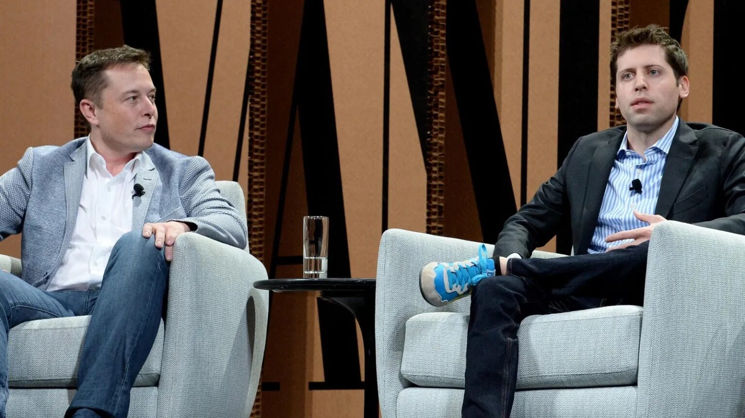 Dois homens sentados em poltronas cinzas num palco, conversando, com um copo d'água sobre a mesa lateral e microfones de lapela