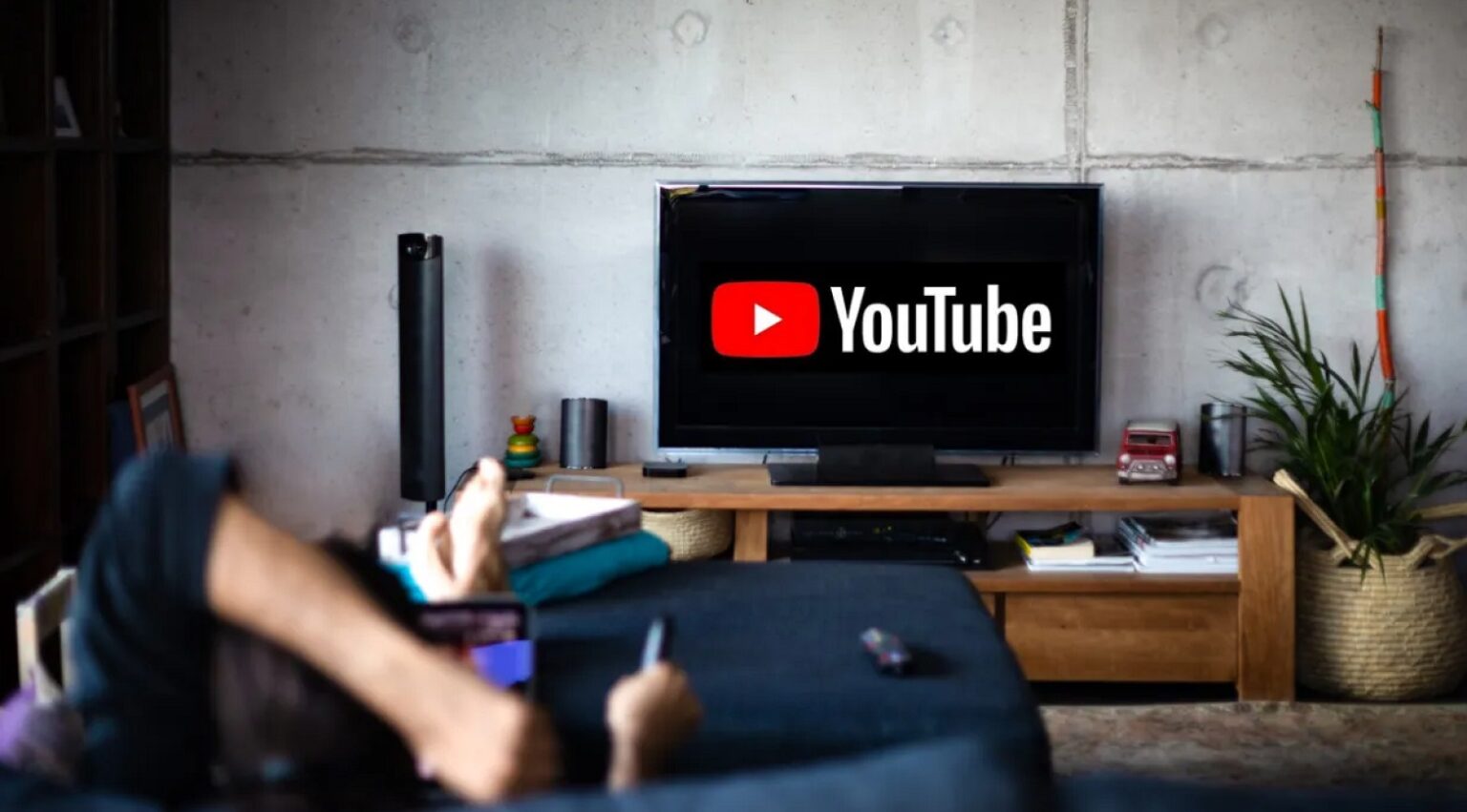 Pessoa deitada no sofá usando celular, com TV de sala exibindo a logo do YouTube em ambiente doméstico