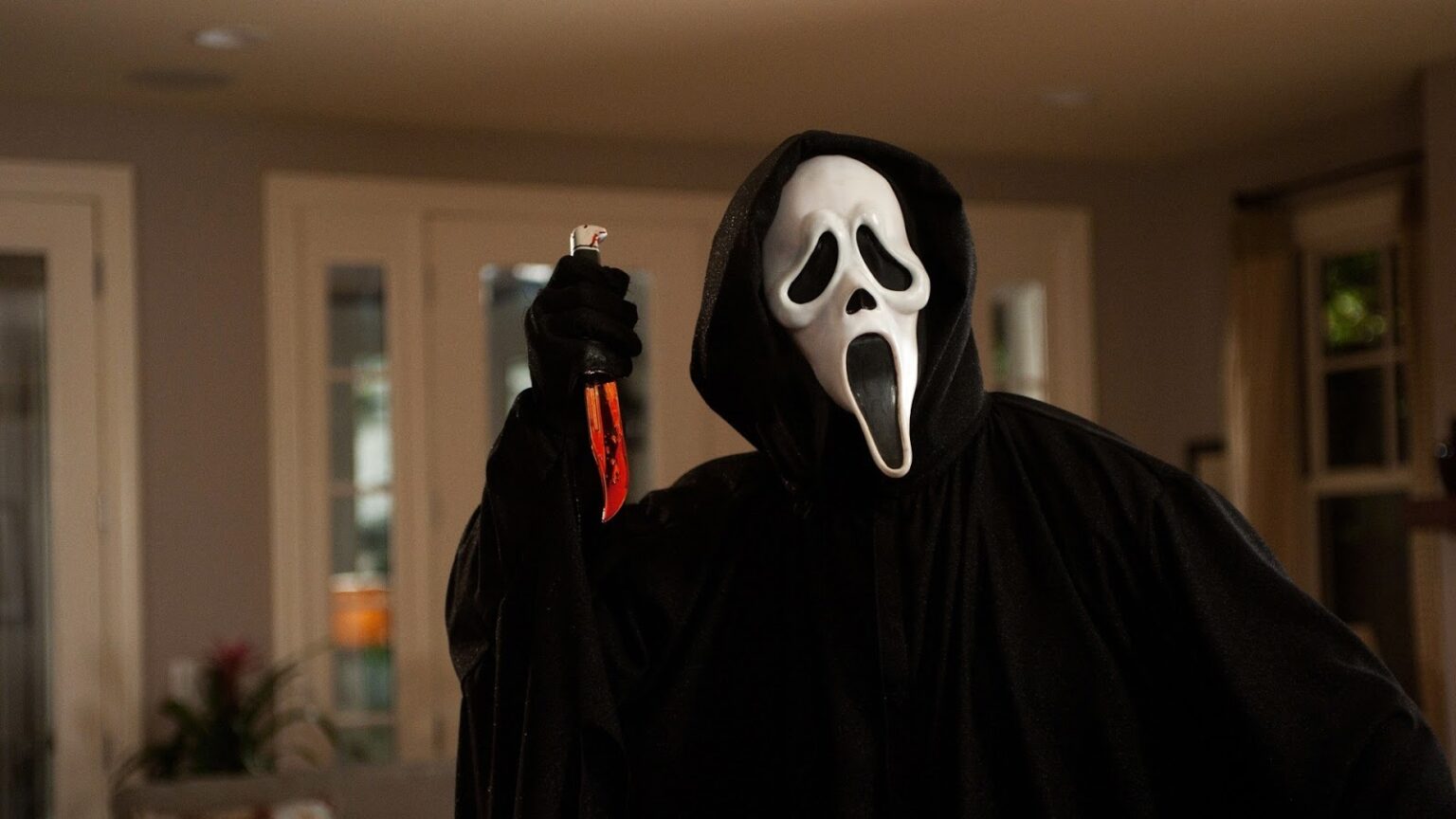 Figura mascarada de manto preto e máscara branca de rosto gritando segura uma faca ensanguentada em uma sala de estar, cena de filme de terror slasher