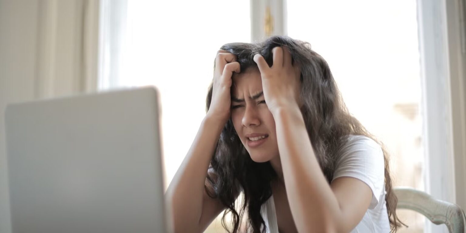 Mulher de cabelos castanhos com as mãos na cabeça e expressão de frustração diante de um notebook prateado aberto na mesa