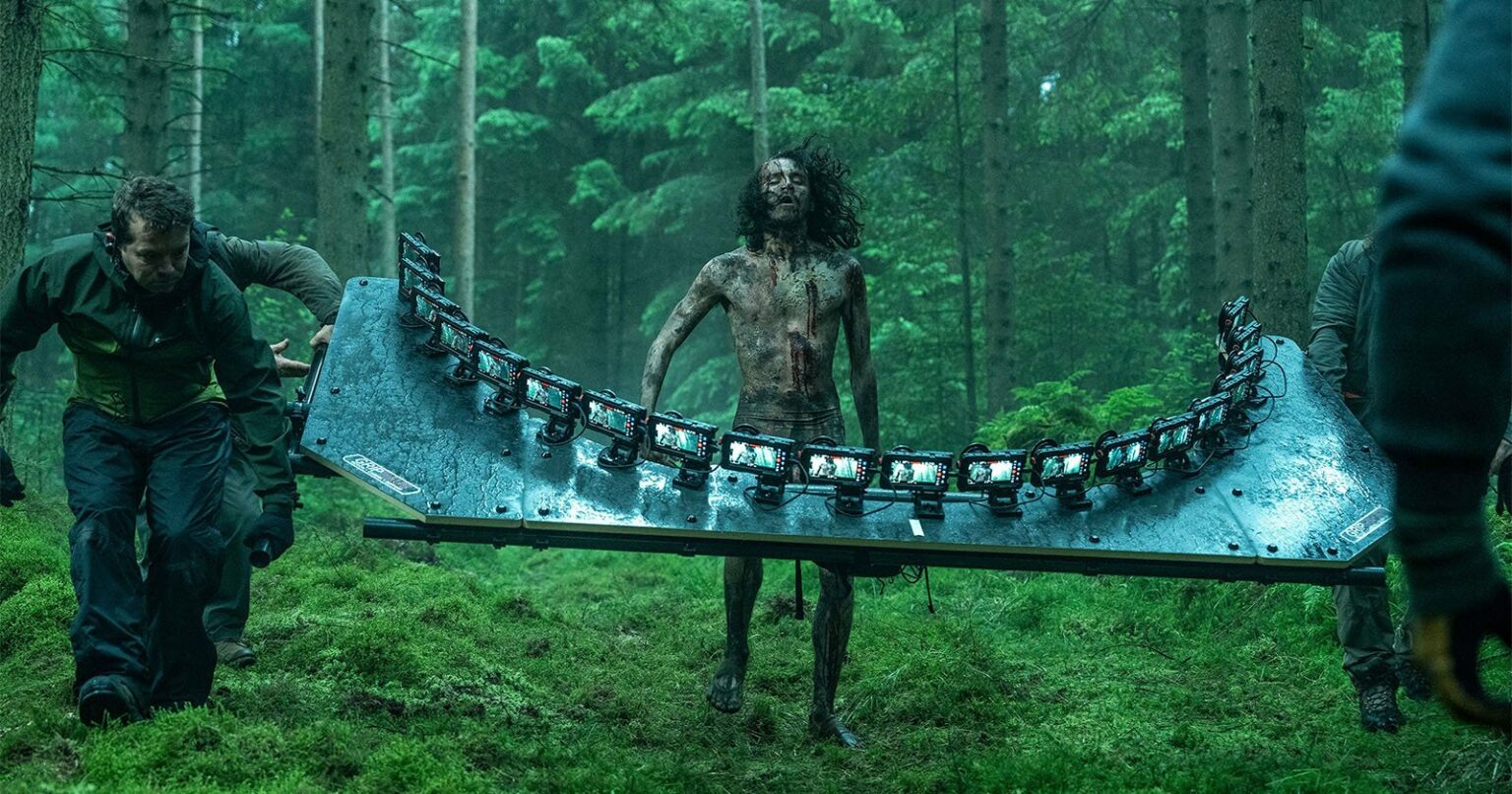 Homens em uma floresta carregando uma tábua metálica sobre a qual há fileiras de luzes, enquanto um homem de corpo sujo e cabelos longos caminha no centro