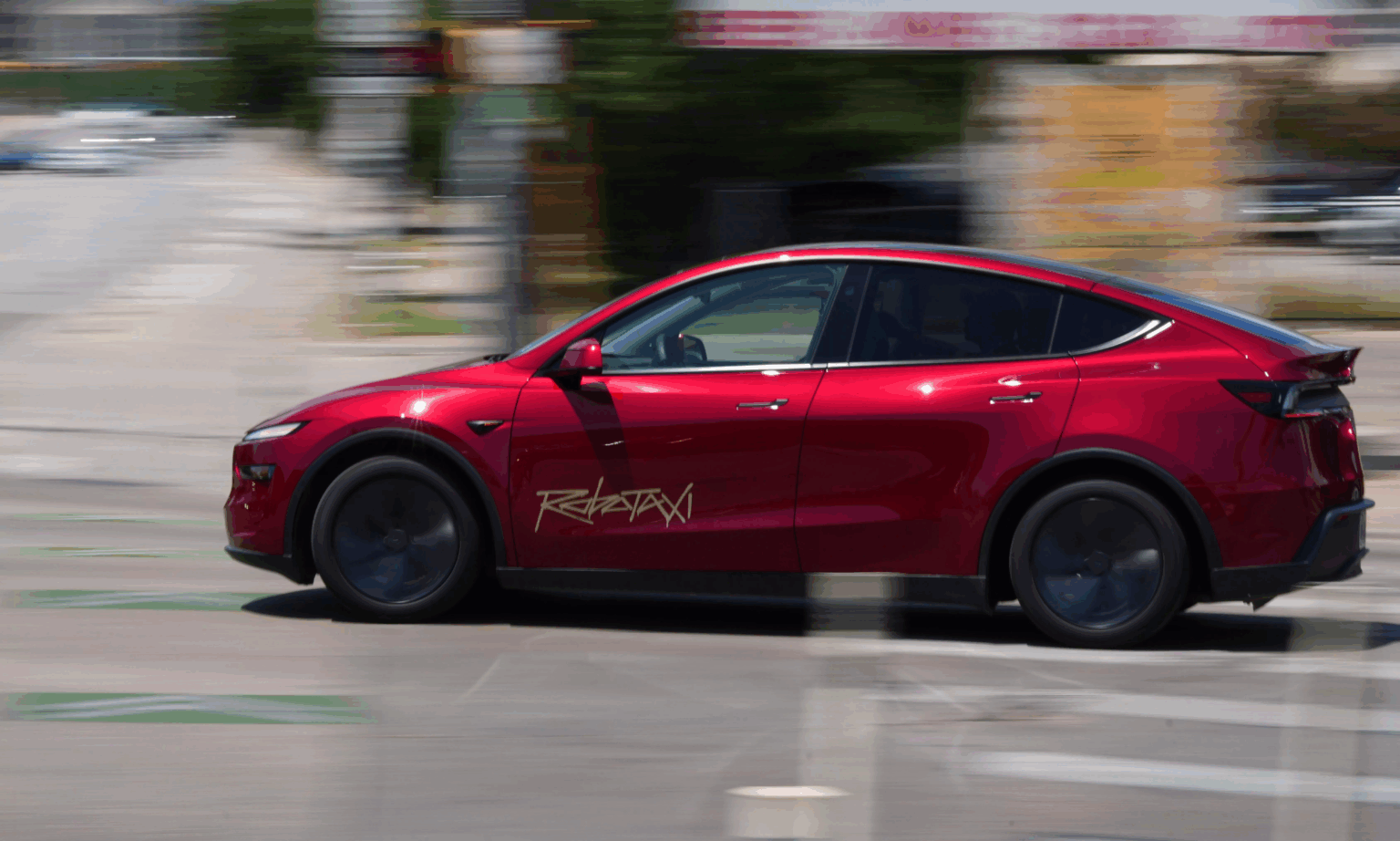 Carro elétrico vermelho em movimento, modelo Tesla Model Y, com o adesivo "Robotaxi" dourado na porta lateral, em uma rua urbana
