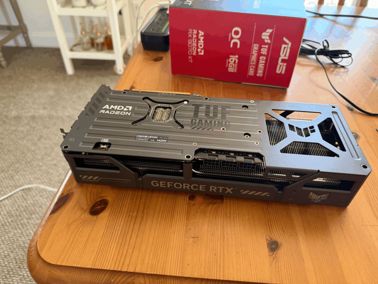 Placa de vídeo ASUS TUF Gaming sobre mesa de madeira, com "AMD Radeon" e "GeForce RTX" impressos no mesmo cooler, e caixa vermelha da Radeon RX 9070 XT ao fundo