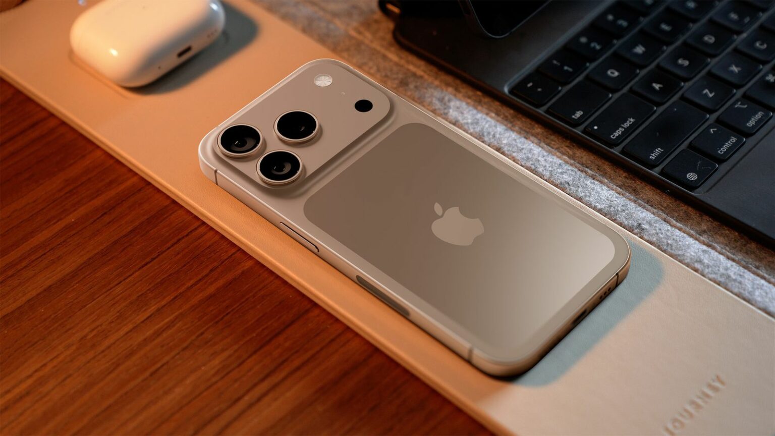 iPhone 17 Pro na cor dourada, com câmera traseira tripla e logo da Apple, deitado sobre um mouse pad ao lado de um teclado e de um case de AirPods