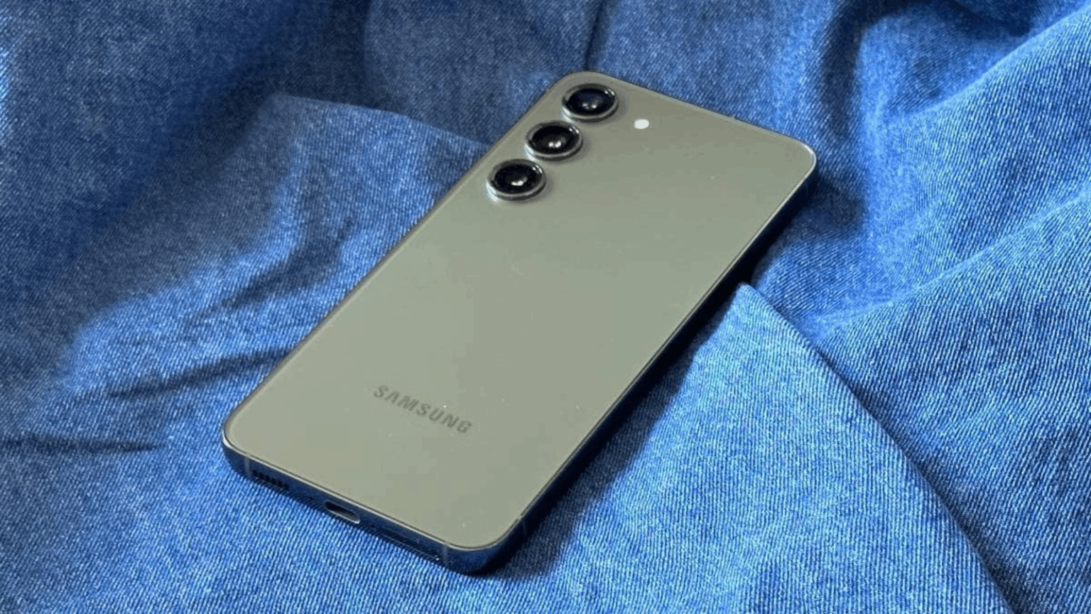 Smartphone Samsung Galaxy verde-acinzentado com três câmeras traseiras, deitado sobre um tecido azul de jeans amassado