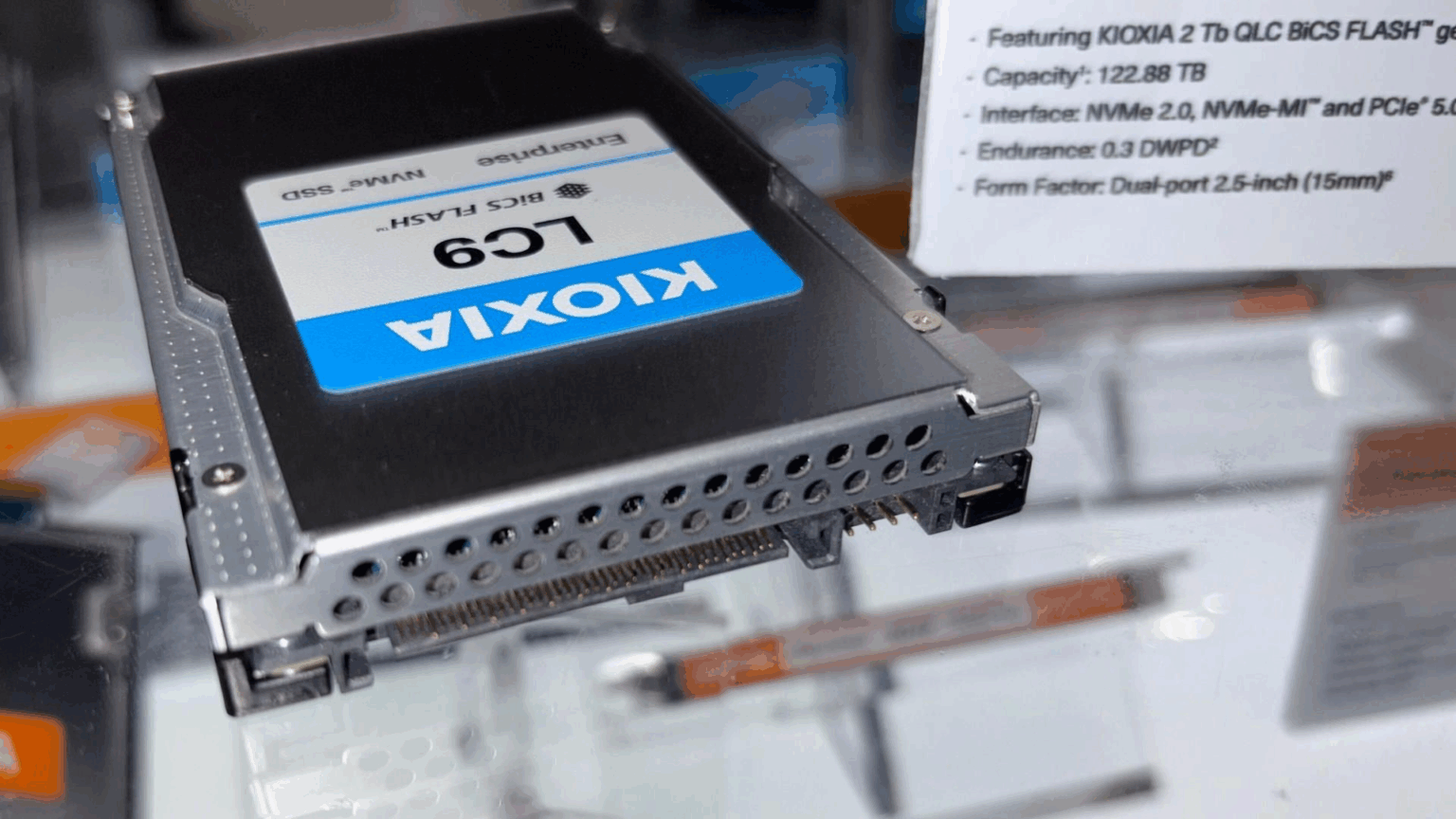 SSD Kioxia LC9 Enterprise NVMe de 122,88 TB em exibição em feira, com etiqueta azul da marca e ficha técnica ao fundo