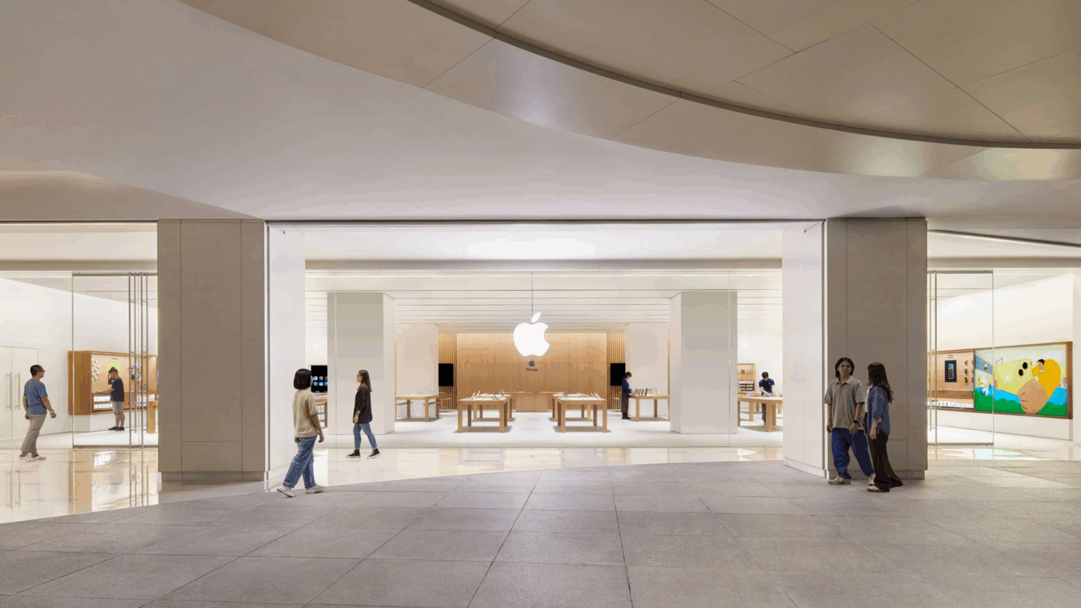 Fachada interna de loja Apple com logo iluminado ao fundo, mesas de produtos expostas e pessoas caminhando no piso claro