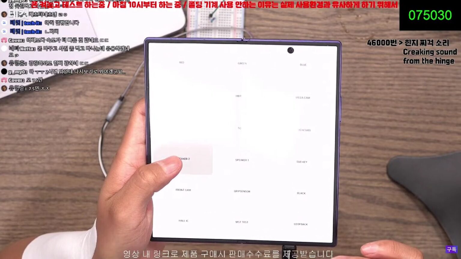Captura de transmissão ao vivo mostrando mãos segurando um Galaxy Z Fold aberto, com tela de teste de componentes e chat de espectadores ao redor