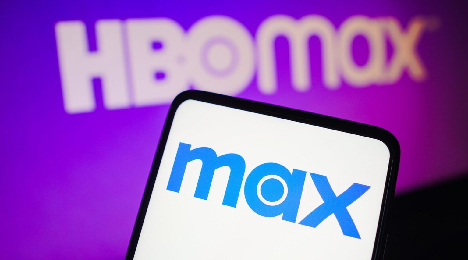 Smartphone exibindo o logo azul da Max em tela branca, com o logo HBO Max em branco ao fundo sobre um gradiente roxo