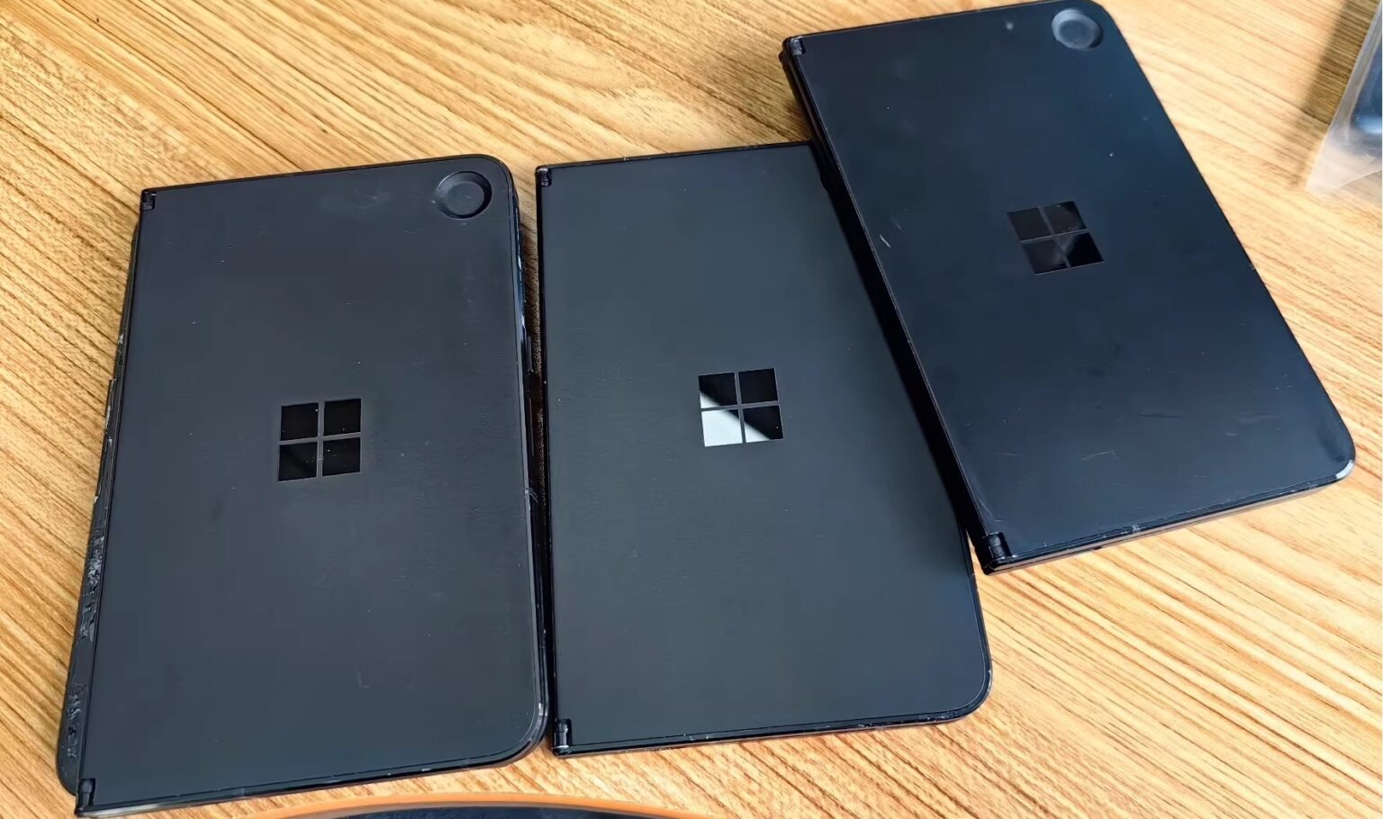 Três dispositivos dobráveis pretos da Microsoft, com logo Windows, dispostos sobre uma mesa de madeira