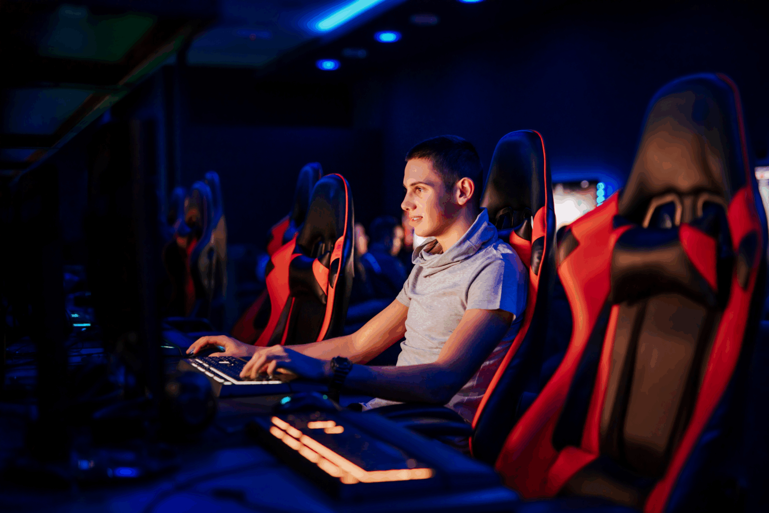 Jovem jogando em computador com teclado iluminado, sentado em cadeira gamer vermelha e preta em ambiente escuro com luzes azuis