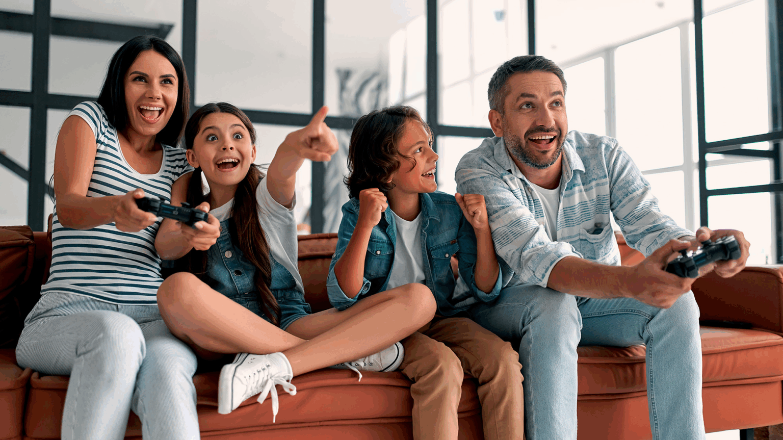 Família animada jogando videogame com controles no sofá, uma mãe, duas crianças e um pai comemorando juntos na sala