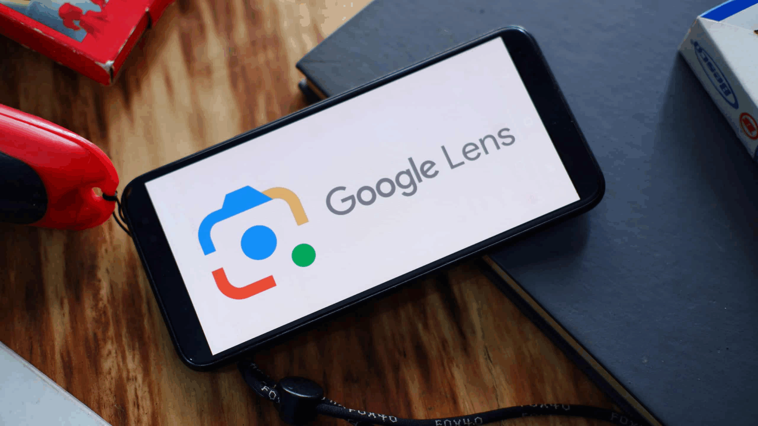 Smartphone exibindo o logotipo do Google Lens sobre uma mesa de madeira, ao lado de um caderno e um objeto vermelho