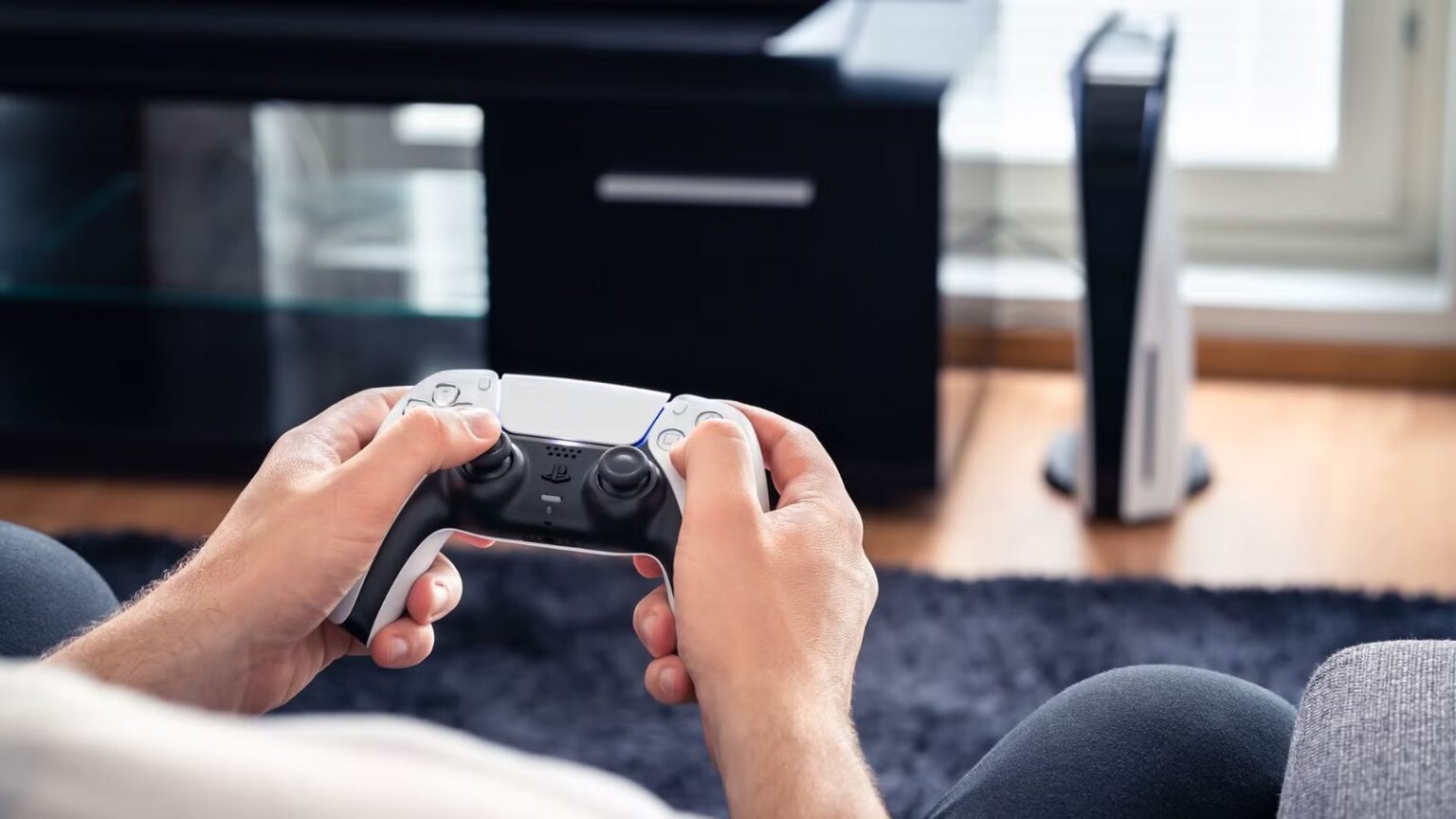 Mãos segurando um controle DualSense branco e preto de PlayStation 5 em uma sala de estar, com o console PS5 ao fundo